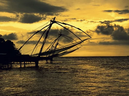 Chinese fishing nets at Fort Kochi Kerala during dramatic sunset, featuring silhouetted cantilevers over Arabian Sea, cloudy golden sky, perfect iconic Kochi tour package.