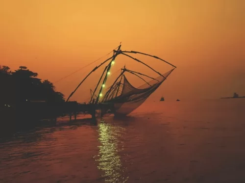 Illuminated Chinese fishing nets at Fort Kochi Kerala during orange sunset glow, featuring cantilever nets over calm sea, silhouetted palms, perfect romantic Kochi tour package. 