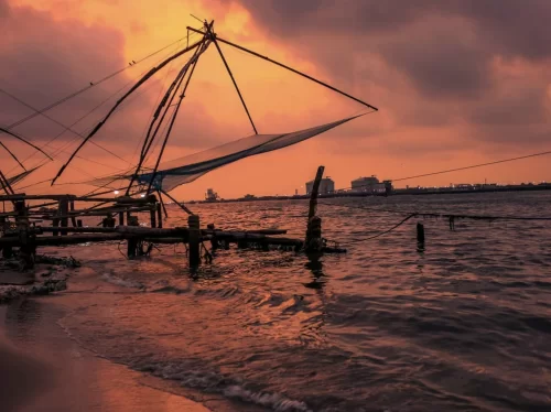 Chinese fishing nets silhouette at Fort Kochi Kerala during fiery orange sunset, featuring dramatic cloudy sky, cantilever nets over Vembanad Lake shore, perfect romantic Kochi tour package.