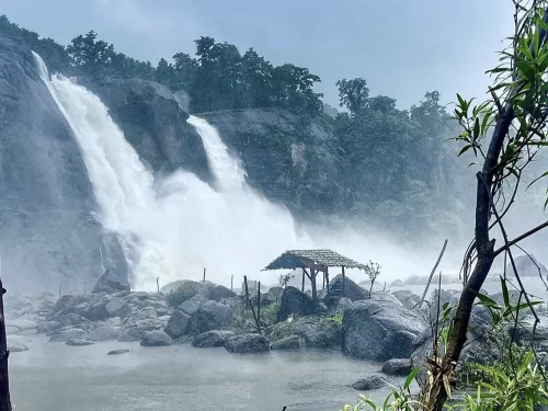 Athirappilly Waterfalls Thrissur Chalakudy Kerala India dramatic tall waterfall pouring rocky cliff lush rainforest overcast rainy skies foreground bamboo hut rocks shallow pools green foliage, perfect Kerala nature photography tour package.