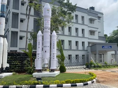 Thiruvananthapuram Kerala ISRO Space Centre entrance four white rocket models GSLV PSLV tricolor flag India Pro Hindi text grey building palm trees green fence lawn landscaping cloudy sky, space museum educational tour package