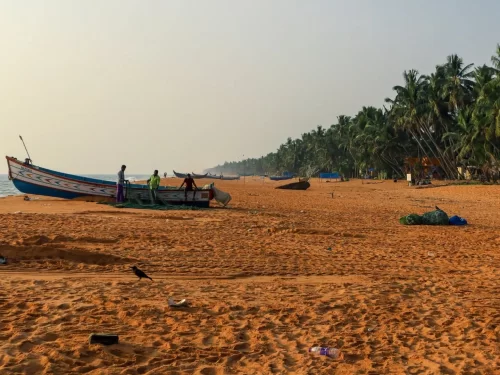 Thiruvananthapuram Kerala Thumba Beach golden sand traditional blue painted fishing boats nets fishermen beach huts coconut palm trees hazy sunset sky serene coastal village scene tour package. 