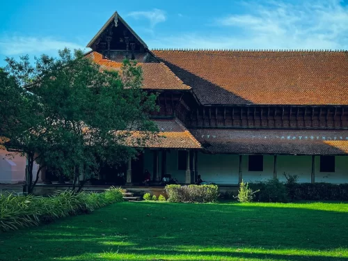 Kuthiramalika Palace Thiruvananthapuram Kerala elegant facade with terracotta roofs, carved wooden pillars, shaded verandas and well-kept green courtyard under blue sky, perfect India tour package. 