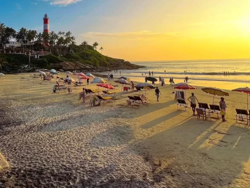 Golden hour at Kovalam Beach Kerala with iconic red-white lighthouse on cliff, colorful beach umbrellas, loungers, tourists strolling golden sand and Arabian Sea waves, perfect India tour package. 