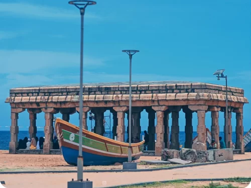 Iconic Arattu Mandapam stone pavilion with tall pillars on Shanghumugham Beach Thiruvananthapuram featuring colorful traditional Kerala fishing boat docked nearby surrounded by modern street lamps people under vibrant blue skies Arabian Sea, perfect Keral