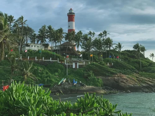 Kovalam Vizhinjam Lighthouse red white striped tower hilltop lush green vegetation palm trees flags buildings surfers ocean waves rocky coast Kerala India scenic landmark tourism photography cloudy sky.