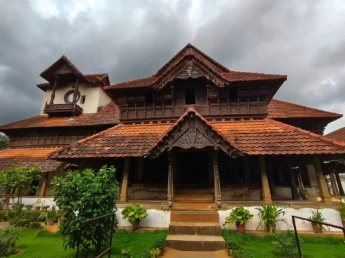 Front courtyard of Padmanabhapuram Palace during cloudy daytime, featuring traditional Kerala wooden architecture, tiled roofs and green garden, perfect Kanyakumari heritage tour package