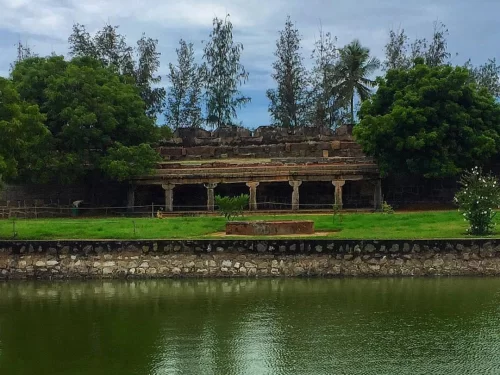 Ancient pillared mandapa ruins amid casuarina trees and lush greenery beside serene pond at Shore Temple complex in Mahabalipuram, Tamil Nadu, perfect Tamil Nadu tour package.