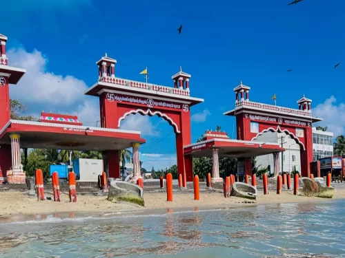 Agni Teertham Rameshwaram Ramanathapuram Tamil Nadu India majestic red triple arched entrance gate Tamil inscription flags atop beachfront shallow turquoise waters sandy shore coconut palms clear blue skies birds flying, perfect South India temple coastal
