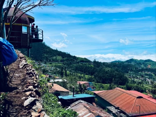 Ketti Valley View Point Ooty Nilgiris Tamil Nadu India closeup featuring maroon dome glass-walled observation deck balcony with tourists overlooking clear blue skies green hills terraced fields colorful tin roofed village homes stone walls blue tarp cover
