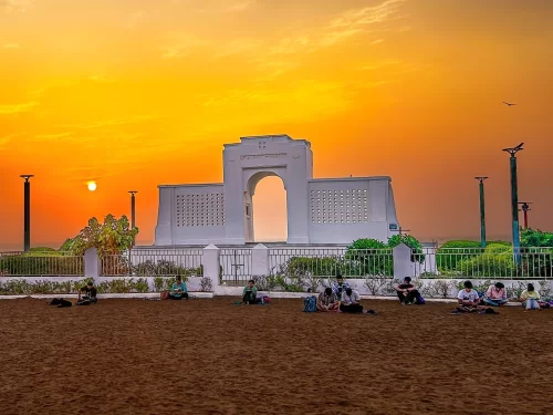 Karl Schmidt Memorial at Besant Nagar Beach during golden hour sunset, featuring white Art Deco archway, silhouetted palms, relaxed visitors on sands, perfect coastal serenity with Chennai beach tour package.
