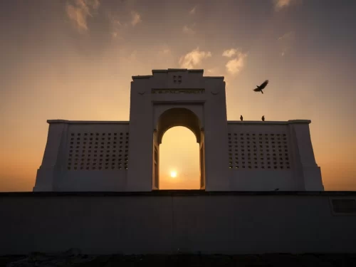 Karl Schmidt Memorial at Besant Nagar Beach during dramatic sunset silhouette, featuring white lattice archway framing sun, flying bird, cloudy skies, perfect romantic coastal experience with Chennai beach tour package.