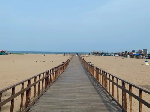 Wooden pier walk at Besant Nagar Beach during clear daytime, featuring endless golden sands, distant fishing boats, sea horizon, perfect adventure stroll with Chennai beach tour package.