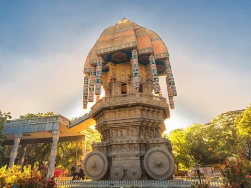 Golden hour sunlight Valluvar Kottam Chennai stone chariot trees flowers blue hazy sky, featuring terracotta dome colorful drapes detailed carvings wheels entrance, perfect majestic Tamil literary monument Chennai tour package.