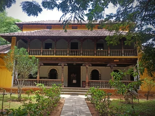 Traditional two-storey heritage house at DakshinaChitra Heritage Museum, featuring wooden balcony, tiled roof, yellow walls, and green garden pathway showcasing classic South Indian architecture.