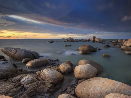 Granite boulders at Samudra Beach during dramatic sunset, featuring massive rocks, turquoise waves, cloudy skies, perfect romantic experience with Kovalam Samudra Beach tour package.