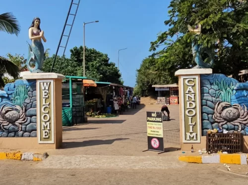 Iconic mermaid entrance gate at Candolim Beach Goa during sunny day, featuring colorful Welcome Candolim signage crabs parking sign shops trees, perfect beach access experience with Candolim Beach Goa tour package. 