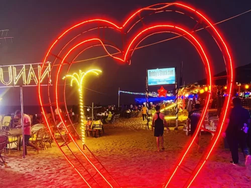 Glowing red heart arch at night at Candolim Beach Goa during vibrant evening, featuring Kulaman signage illuminated shacks seating crowds, perfect romantic nightlife experience with Candolim Beach Goa tour package.