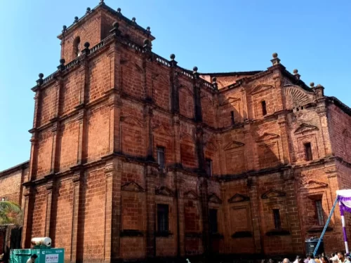 Basilica of Bom Jesus Old Goa during sunny day, featuring ornate red laterite facade towers balustrades, tourists construction scaffolding flags palm tree foreground, perfect UNESCO heritage Portuguese baroque church experience with South Goa old Goa tour