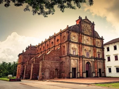 Sepia-toned Basilica of Bom Jesus Old Goa under cloudy sky with tree overhang, featuring weathered red laterite facade intricate carvings towers, adjacent white Professão de Fé church palms grass foreground, perfect vintage UNESCO Portuguese baroque herit