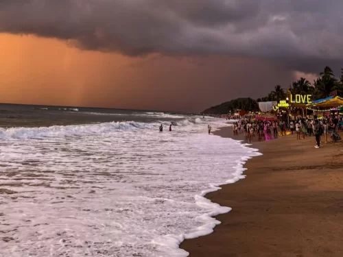 Goa Anjuna Beach golden hour dramatic orange purple stormy clouds yellow LOVE beach shack sign crowd tourists colorful sarees people walking swimming sandy shore ocean waves palm trees coastal party vibe sunset