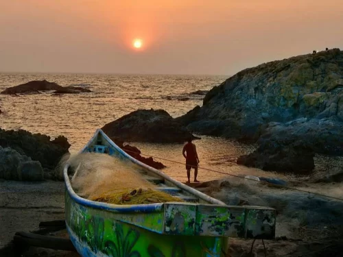 Iconic Vagator Beach Goa at sunset with fisherman silhouette beside colorful traditional boat on rocky shore, Arabian Sea waves and dramatic orange sky, perfect romantic beach photography with North Goa tour package.