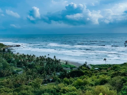 Epic Vagator Beach Goa cliff top vista with crashing blue waves, sandy cove, palm groves, beach huts under partly cloudy skies, perfect thrilling North Goa beach adventure with tour package. 