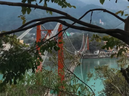 Laxman Jhula new suspension bridge at Rishikesh over Ganga River, through trees featuring red towers cables Neelkanth Temple hilltop buildings, perfect spiritual experience Uttarakhand tour package.