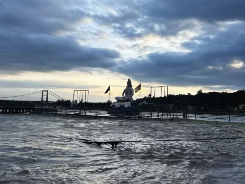 Panoramic view colossal Shiva statue with fluttering saffron flags overlooking swirling Ganga River Rishikesh during dramatic cloudy sunset, suspension bridge poles distant hills townscape water flow railing foreground, perfect Uttarakhand tour package.