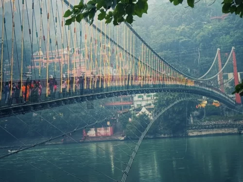 Laxman Jhula suspension bridge at Rishikesh during foggy morning, featuring yellow cables temple buildings Ganga River trees walkers, perfect spiritual experience Uttarakhand tour package.