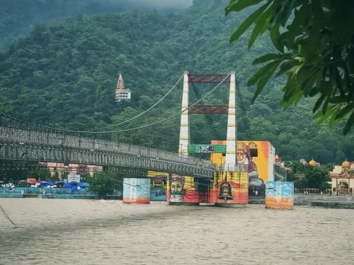 Janki Setu suspension bridge in Rishikesh wide view showing iron towers cables colorful murals pillars Ganges river lush green hills distant temples trees backdrop, perfect spiritual adventure Uttarakhand tour package.