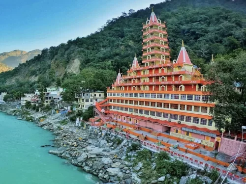 Wide-angle view Trayambakeshwar Temple Rishikesh along Ganga riverbank during clear day, showcasing orange multi-story pagoda towers forested mountains riverside buildings rocks greenery, perfect spiritual riverside heritage Uttarakhand tour package.