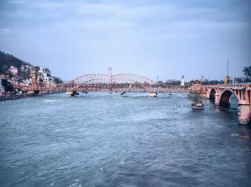 Har Ki Pauri Haridwar iconic pink arched Lakshman Jhula bridge over Ganga river temples boats hills cloudy sky, perfect pilgrimage riverside view, Uttarakhand tour packages.