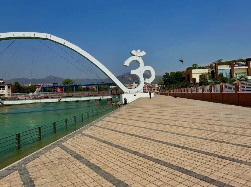 Har Ki Pauri Haridwar iconic white Om symbol on modern curved pedestrian bridge over Ganga river mountains blue sky green trees, perfect spiritual riverside walk, Uttarakhand tour packages.