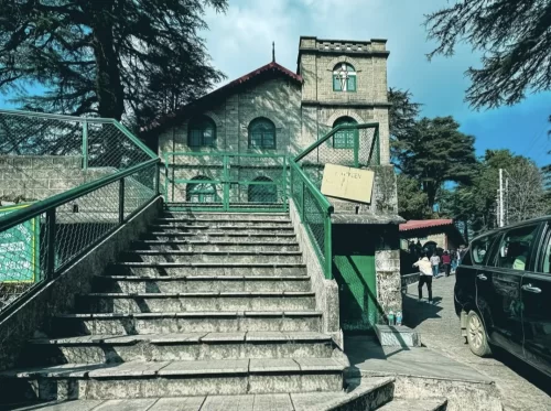 St Paul's Church entrance at Landour during partly cloudy day, featuring stone tower, green railings, stairs and deodar trees, perfect romantic Mussoorie tour package.
