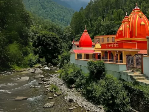 Kainchi Dham temple amidst lush greenery during sunny day, featuring vibrant orange shikhara domes yellow red buildings forested Himalayan hills flowing rocky river compound wall, perfect spiritual cultural experience Uttarakhand tour packages.