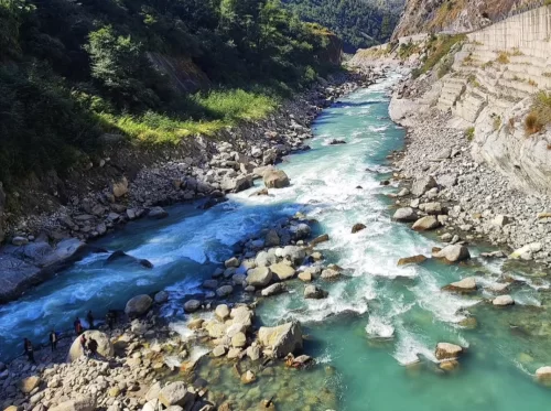 Vishnuprayag sacred confluence of turquoise Alaknanda and Dhauliganga rivers amid lush green forested hills and rocky cliffs, with sunlight sparkling on rushing waters and distant people, perfect Uttarakhand tour package.