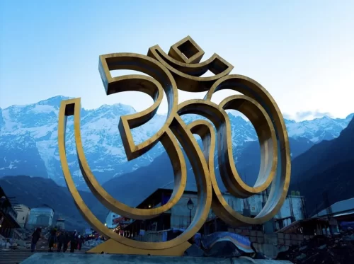 Giant golden Om symbol at Kedarnath during sunny day, featuring snowy Himalayan peaks blue skies visitors foreground, perfect spiritual Uttarakhand tour packages.