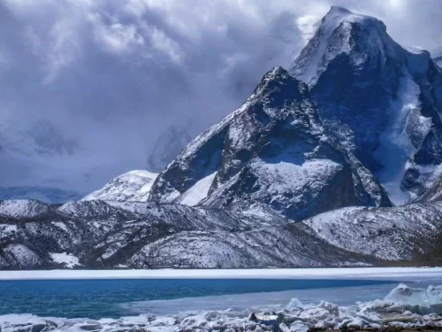 Gurudongmar Lake reflection panorama at North Sikkim during sunny day, featuring snow peaks prayer flags turquoise waters rocky foreground, perfect high-altitude adventure Sikkim tour package.