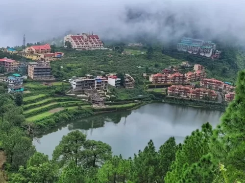 Khurpa Tal Nainital misty lake panorama during foggy day, featuring red roofed resorts terraced fields pine hills mist backdrop, perfect nature romantic experience Uttarakhand tour packages.