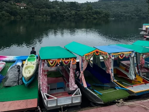 Decorated shikara boats docked at Naukuchiatal Lake Nainital during cloudy daytime, featuring colorful canopies and forested hills, perfect romantic experience Uttarakhand tour package.