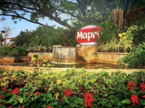Mapro Garden at Mahabaleshwar during sunny day, featuring Mapro logo sign, cascading waterfall, tropical plants, flower beds, perfect family experience Maharashtra tour packages.