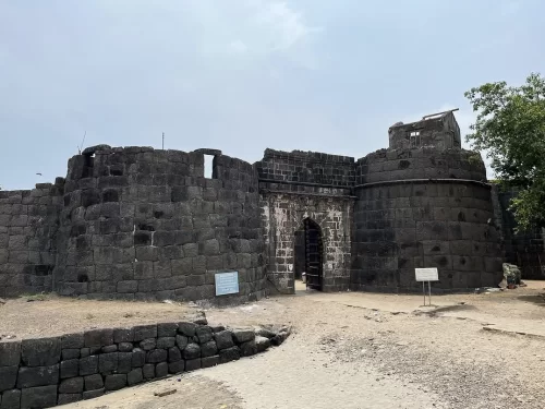 Ground-level view of Kolaba Fort’s massive black stone walls and arched entrance gate, with rounded bastions, weathered masonry, and open sandy foreground under a clear sky.