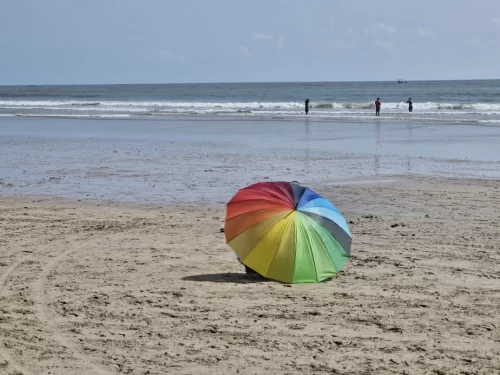 Vibrant rainbow umbrella on Kashid Beach during overcast daytime, featuring endless golden sands, gentle waves, distant figures, perfect beach relaxation experience with Konkan coast tour package.