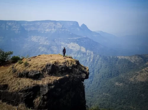 Louisa Point Matheran solo trekker cliff edge golden grass distant plateau mountains valleys hazy skies Western Ghats, perfect Maharashtra hill station dramatic viewpoint adventure, Maharashtra tour packages.