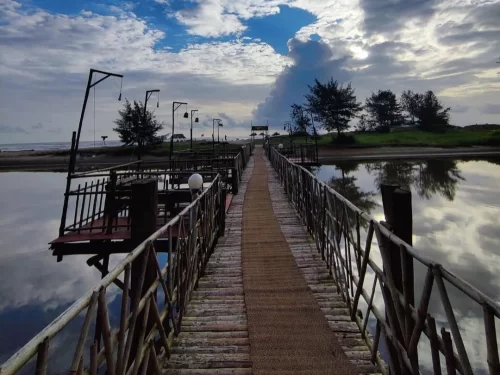 Long exposure view of Ashwem Beach wooden jetty walkway near Mandrem Beach North Goa during dramatic cloudy sunset, featuring calm reflective lagoon palm trees beach umbrellas lamps, perfect serene nature experience with Mandrem Ashwem Beach Goa tour pack
