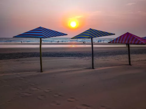 Golden hour silhouette of colorful striped beach umbrellas on sandy shore Mandrem Beach North Goa with setting sun Arabian Sea waves distant people, perfect romantic sunset experience with Mandrem Beach Goa tour package.