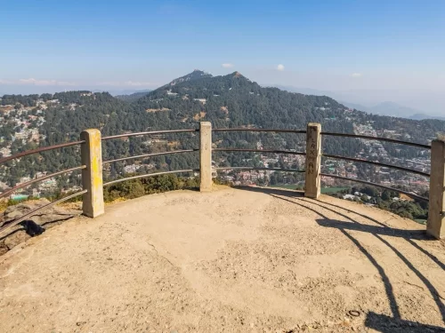 Tiffin Top Nainital elevated concrete viewpoint rusty yellow railings overlooking pine-clad hills terraced town distant snow peaks hazy blue skies clear weather, perfect Uttarakhand tour packages.