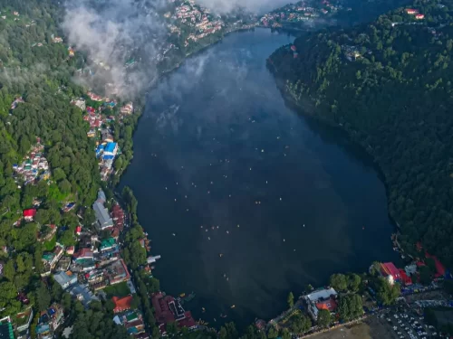 Aerial misty cloudy drone view of Naini Lake Nainital, featuring pear-shaped dark waters, scattered boats, colorful hillside town and forested hills, perfect adventure experience Uttarakhand tour package.