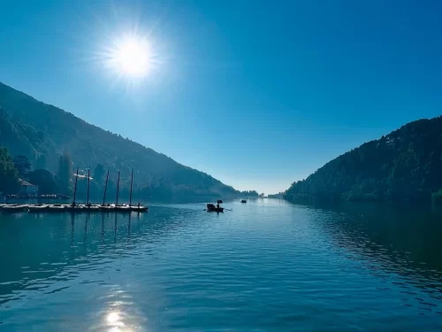 Sunny daytime scene on Naini Lake Nainital, featuring docked masted boats, rowboat and distant mountains, perfect adventure experience Uttarakhand tour package.